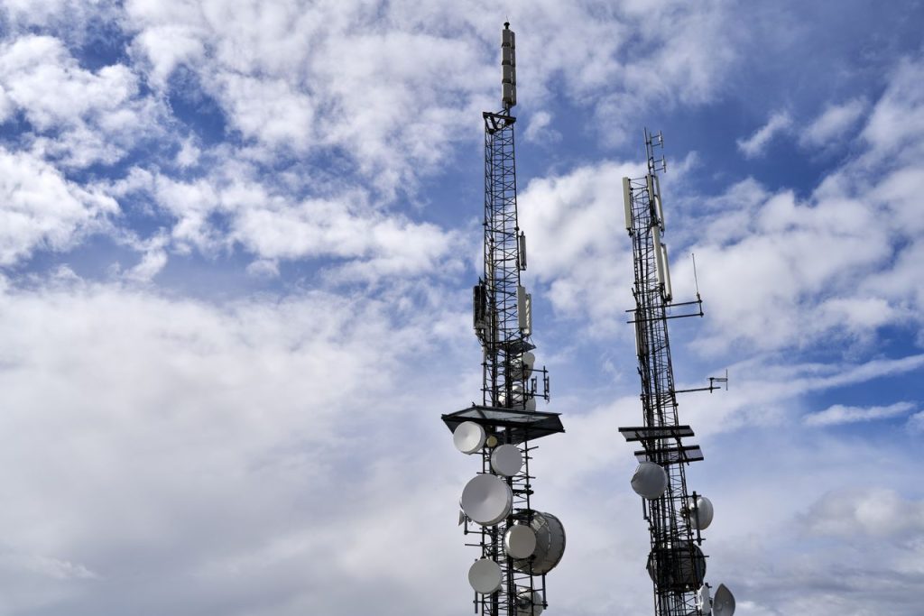 Service Telecommunication towers with antennas and dishes under a bright cloudy sky.